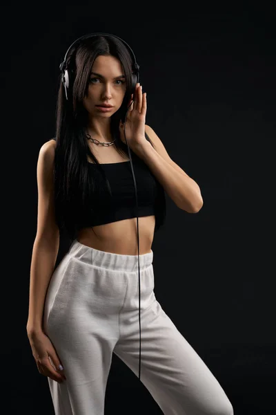 Woman wearing fashion sport clothes listening music with headphones inside. Front view of pretty female model in black top posing with earphones, isolated on black background. Concept of fashion.