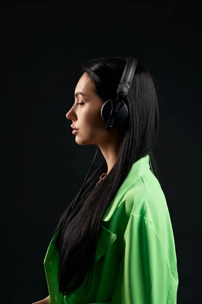 Calm girl in green shirt listening to music with headphones inside. Side view of serious woman with dark hair and large over-ear headset, isolated on black studio background. Concept of posing.