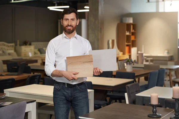 Front view of brunette male with beard standing in furniture store. Handsome designer, architect, seller showing wooden samples, smiling, looking at camear. Concept of furniture.