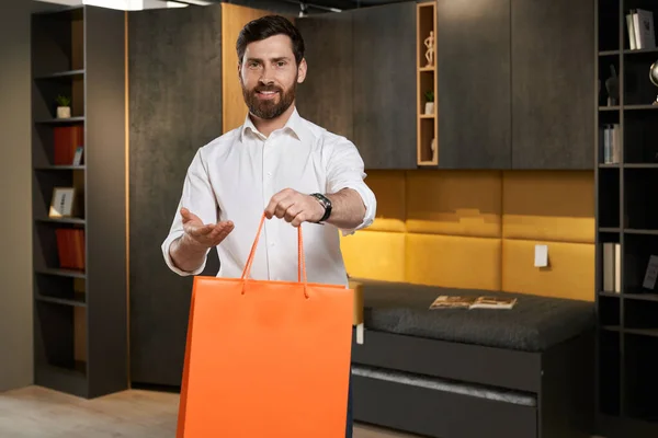 Front view of handsome consumer standing, holding, showing giving bag in furniture store. Brunette man smiling, looking at camera, happy, glad, satisfied. Concept of shopping.