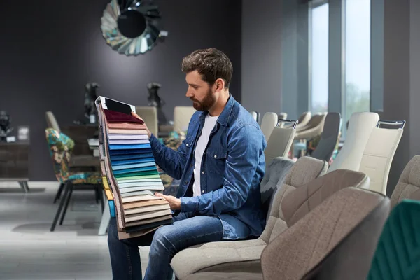 Side view of young brunette male thinking, choosing fabric in furniture store. Handsome boy sitting on chair, holding palette with colors, looking, touching. Concept of shopping.