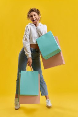 Beyaz kapüşonlu mutlu kadın alışveriş torbaları tutuyor, stüdyoda poz veriyor. Paketleri olan, gülümseyen, utangaç bir şekilde el ele tutuşan, turuncu arka planda izole edilmiş güzel bir model. Alışveriş kavramı.