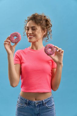 Gözleri kapalı neşeli bir kadın, elinde iki renkli çörek, stüdyoda gülümsüyor. Kıvırcık saçlı, pembe serpiştirilmiş donutları olan, gök mavisi arka planda izole bir kadın. Eğlence anlayışı.