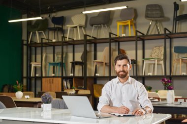 Gülümseyen yönetici uzaktan çalışırken dizüstü bilgisayarla masada oturuyor. Arka planda sandalyeli raflara karşı yazarken kameraya bakan kendine güvenen perakendecinin ön görüntüsü. Çalışma alanı kavramı. 