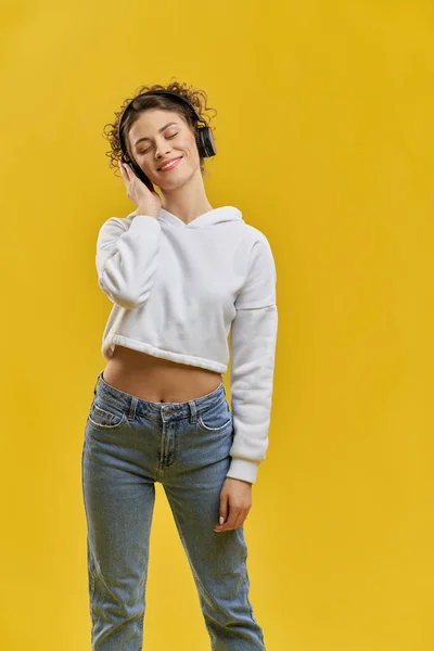 Happy woman with closed eyes standing and listening to music indoor. Front view of focused pretty girl wearing headphones, enjoying music, isolated on orange studio background. Concept of music fan.