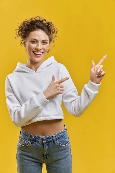 Attractive woman pointing with fingers to one side, looking at camera indoors. Portrait of joyful girl pointing away, smiling, isolated on yellow studio background, copy space. Concept of gesturing.