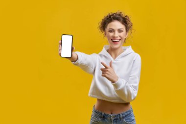Elinde akıllı telefonla poz veren çekici genç modelin ön görüntüsü. Güzel genç bayan ayakta, ekranı gösteriyor, gülümsüyor, kameraya bakıyor. Sarı stüdyo arka planında izole edilmiş..
