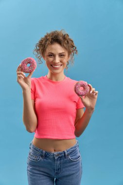 Kıvırcık saçlı, iki donaughts 'ı elleriyle tutan güzel, ince bir kızın yazı tipi. Güzel genç bayan ayakta, gülümsüyor, kameraya bakıyor. Mavi stüdyo arka planında izole edilmiş..