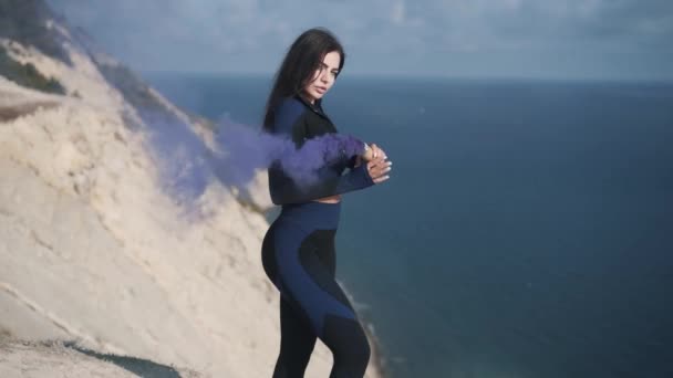 Молода спортивна жінка стоїть на скелі, тримаючи фіолетову димову бомбу — стокове відео