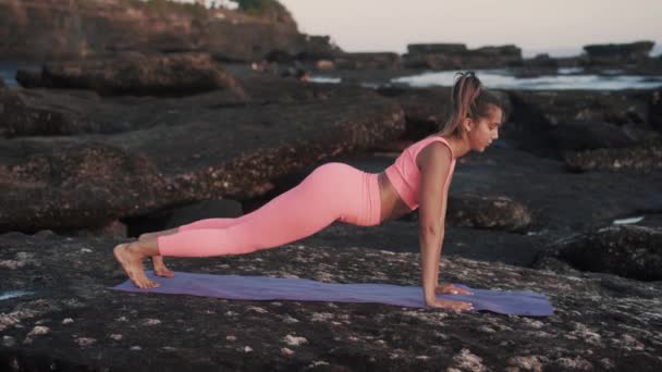 Belle jeune femme faisant des exercices sur tapis de yoga sur le rocher à la plage. — Video