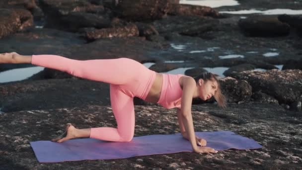 Belle jeune femme faisant des exercices sur tapis de yoga sur le rocher à la plage. — Video