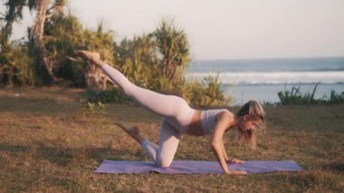 Gün batımında deniz kenarında çalışan genç bir kadın. Kız, dengeyi sağlamak ve göğüs kaslarını güçlendirmek için tek bacak şınavı çekiyor. Spor formda, güçlü ve sağlıklı kalmanıza yardımcı olur. 