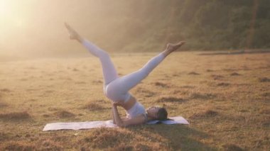 Genç, zinde bir kadın omuz duruşu pozisyonunda bacak egzersizi yapıyor. Pilates antrenmanı sırasında karın kasları ve alt vücut üzerinde çalışıyorum. Gün batımında doğayla birlikte çalış.