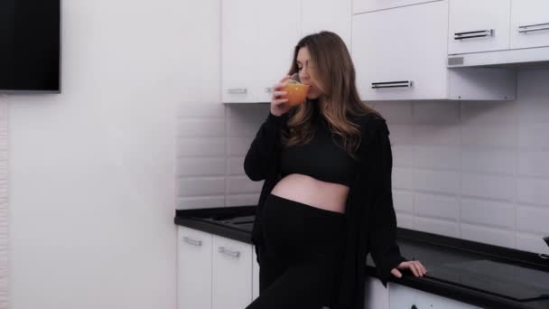 黒い服を着た若い妊婦がキッチンで新鮮なオレンジジュースを楽しんでいます ビタミン柑橘類は 毎日の妊娠食の一部として飲む — ストック動画