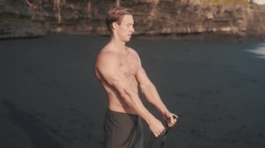 Genç atletik adam kumlu sahilde pazılarını ve trisepslerini çalıştırıyor. Beyaz, mükemmel vücut adamı direniş bandıyla egzersiz yapıyor. Güzellik ve güç kavramı. Yüksek kalite 4k görüntü