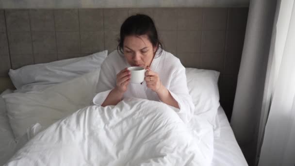 Jonge vrouw in de ochtend liggend in bed en koffie drinkend kijkend naar berichten aan de telefoon. Vrouw wordt wakker en kijkt naar zijn smartphone met een mok warme drank thuis in de slaapkamer. — Stockvideo