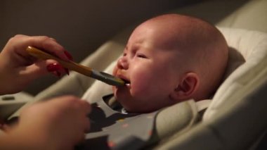 Anne, bebeğe yemek veriyor. Evde şezlongda kaşıktan püre yiyor. Tamamlayıcı bebek beslemesi.