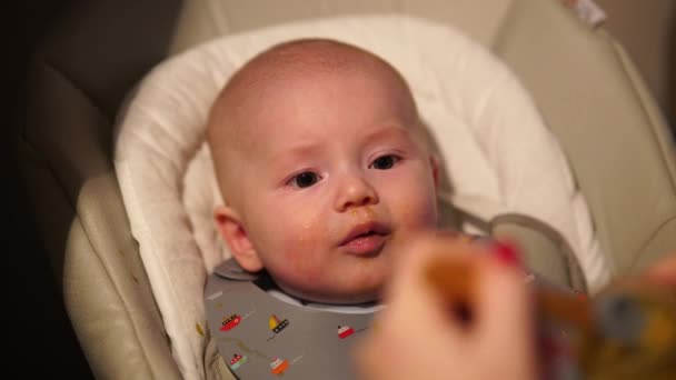 Moeder geeft eten aan baby jongen, puree van lepel in kinderstoel thuis. Aanvullende zuigelingenvoeding. — Stockvideo