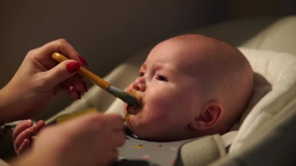 Moeder geeft eten aan baby jongen, puree van lepel in kinderstoel thuis. Aanvullende zuigelingenvoeding. — Stockvideo