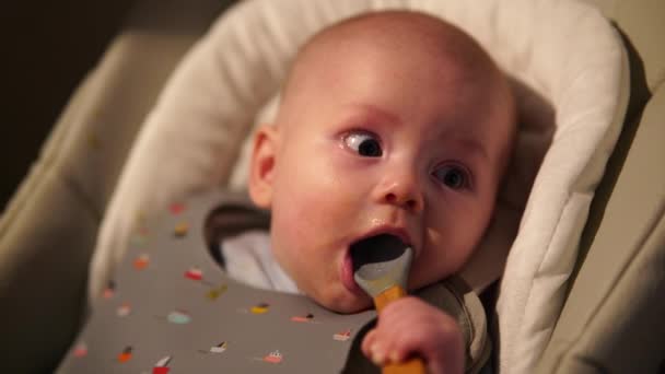 Niño sentado en la silla de alimentación royendo cuchara de goma. Lindo bebé dentición y morder cuchara después de comer. — Vídeos de Stock