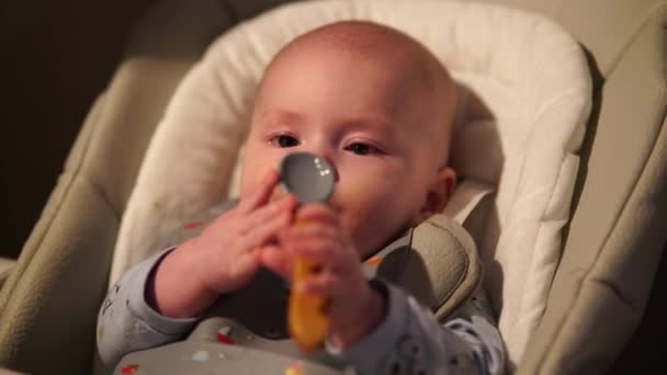 Малыш сидит на кормящем кресле и грызет резиновую ложку. Симпатичные детские зубки и ложки после еды. — стоковое видео