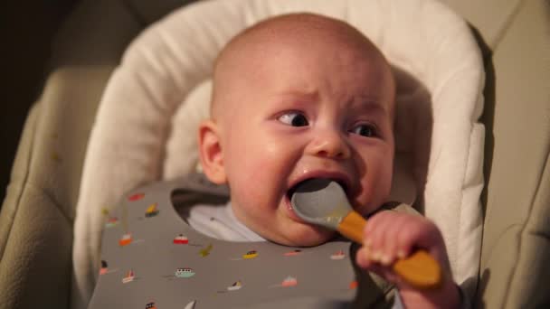 Menino sentado na cadeira de alimentação roendo colher de borracha. Bonito bebê dentição e mordendo colher depois de comer. — Vídeo de Stock