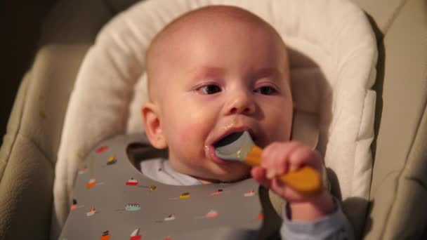 Baby boy sitting on feeding chair gnawing rubber spoon. Cute infant teething and biting spoon after eating. — Vídeo de Stock