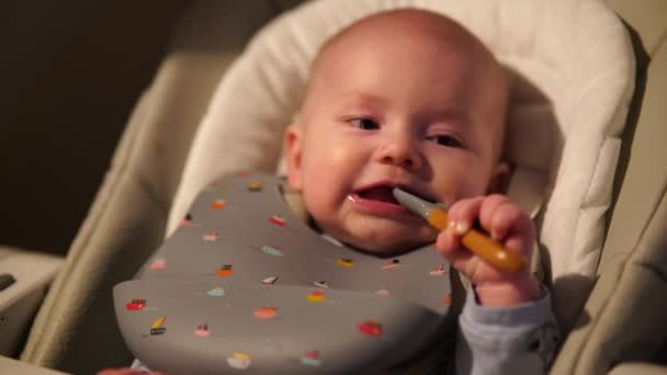 Baby Junge sitzt auf Fütterungsstuhl nagt Gummilöffel. Niedliche Kinderzähne und Beißlöffel nach dem Essen. — Stockvideo