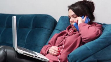 Kanepede oturan kulaklıklı ve dizüstü bilgisayarı olan bir kız. Evdeki koltukta uzaktan çalışma..