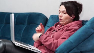Kanepede oturan kulaklıklı ve dizüstü bilgisayarı olan bir kız. Evdeki koltukta uzaktan çalışma..