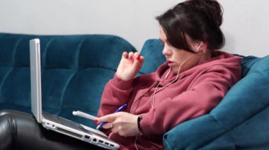 Kanepede oturan kulaklıklı ve dizüstü bilgisayarı olan bir kız. Evdeki koltukta uzaktan çalışma..