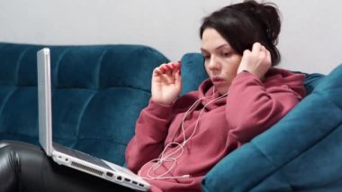 Kanepede oturan kulaklıklı ve dizüstü bilgisayarı olan bir kız. Evdeki koltukta uzaktan çalışma..
