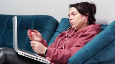 Kanepede oturan kulaklıklı ve dizüstü bilgisayarı olan bir kız. Evdeki koltukta uzaktan çalışma..