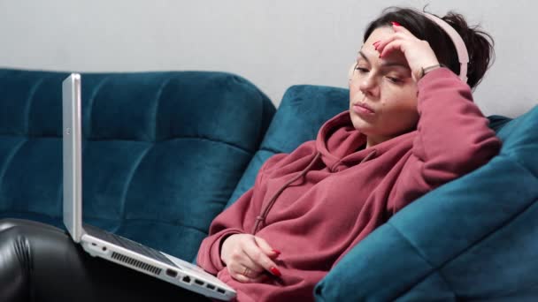 Dívka sedící na pohovce se sluchátky a laptopem. Dálková práce doma na gauči. — Stock video