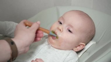 Anne, mutfaktaki sandalyede oturan küçük çocuğunun kaşığıyla besleniyor. Anne bebeği kavanozdan bebek maması ile besliyor..