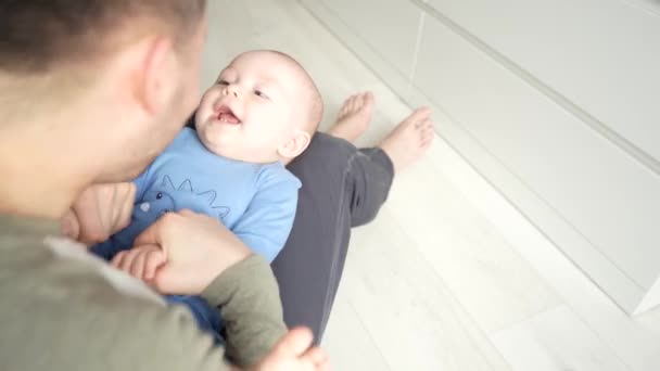 Père joue avec un bébé fils souriant couché sur les genoux à la maison ensemble. — Video