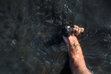 Tuz gölünün suyunda siyah iyileştirici çamur olan bir erkek ayağı. Üst görünüm, seçici odaklanma. Terapi amaçlı çamur üzerinde terapi tatili konsepti.