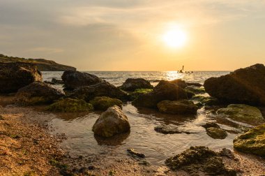 Önünde büyük kayalar olan deniz manzaralı bir akşam. Arka planda, uzakta, banyo yapan insanların siluetleri..