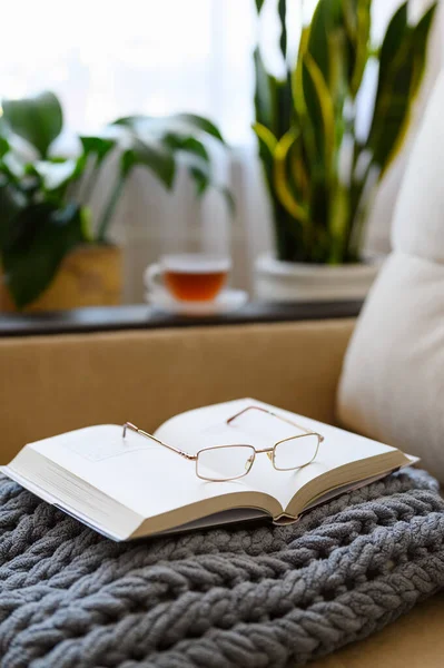 Cozy reading at home on a soft sofa with a cup of tea. An open book with glasses on a gray plaid against the background of a cup of tea and flowers. Vertical format. Selective focus on binding and glasses