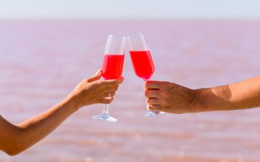 Balayına çıkmış yeni evli bir çift. Ellerinde pembe bir gölün arka planında pembe bir kadın ve erkek kokteyli olan gözlük. Bardağı tutan misk kokulu bir ele odaklan.