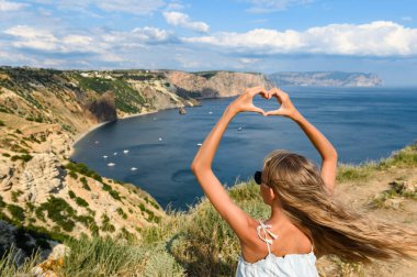 Dağın kenarındaki uzun saçlı bir kız denize bakar ve elleri kalp şeklinde olur. Seyahat ve macera konsepti.