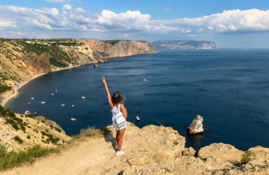 Bir kız denizin arka planına karşı bir dağın tepesinde elleri havada duruyor..