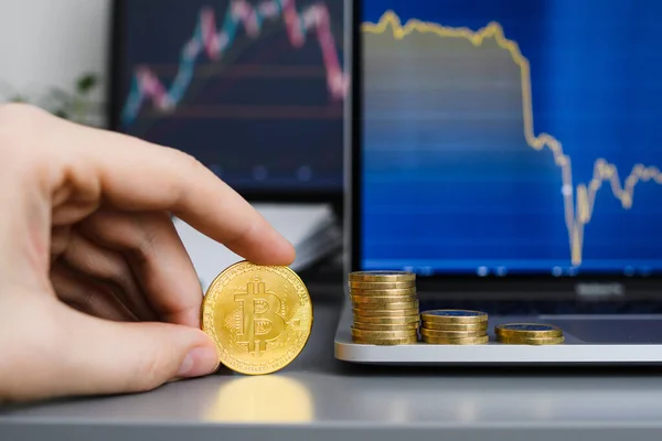 A bitcoin coin in a man's hand against the background of stacks of coins and decreasing graphs on monitors. Concept of falling prices and losses. Selective focus on bitcoin.