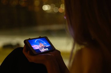 Vladimir, Rusya - Ocak 2022: Geceleri pencerenin önünde bir kız akıllı telefondan Disney filmi izliyor. Logoya seçici odaklanma.