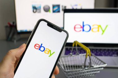 Vladimir, Rusya - Ocak 2022: Bir dizüstü bilgisayar ve alışveriş arabasının arka planına karşı akıllı bir telefonla ebay uygulaması. İnternetten alışveriş yapma fikri. Logoya seçici odaklanma.