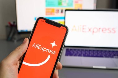 Vladimir, Rusya - Ocak 2022: Bir dizüstü bilgisayarın arka planında elinde AliExpress uygulaması bulunan akıllı telefon. Çin 'den gelen mallar için online alışveriş kavramı. Logoya seçici odaklanma.
