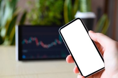 Tabletin arka planında çizelgeler ve bitkilerle birlikte boş beyaz bir ekran bulunan akıllı telefon. Ofisteki işyerinde. Seçici odaklanma. Reklam için şablon.