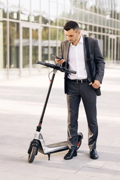 Mężczyzna biznesmen podchodzi do skutera elektrycznego i używa aplikacji na telefon komórkowy. Transport ekologiczny. Człowiek używa skutera elektrycznego jako nowoczesnego środka transportu w mieście — Zdjęcie stockowe