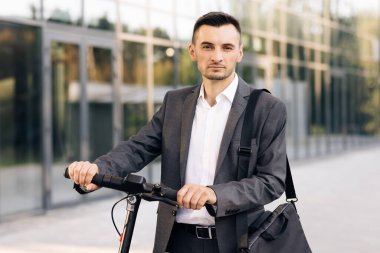 Elektrikli scooterlı kendine güvenen işadamı kameraya bakıyor. E-scooter sürücüsü kişisel çevre aracı kiralıyor. Dışarıdaki araçta stil sahibi adam. Modern kentsel alternatif ulaşım.