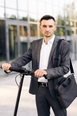 Modern kentsel alternatif ulaşım. Elektrikli scooterlı ve kameraya bakan kendine güvenen iş adamının portresi. Dışarıdaki araçta stil sahibi adam. E-scooter sürücüsü kişisel eko nakliye aracı kiralıyor